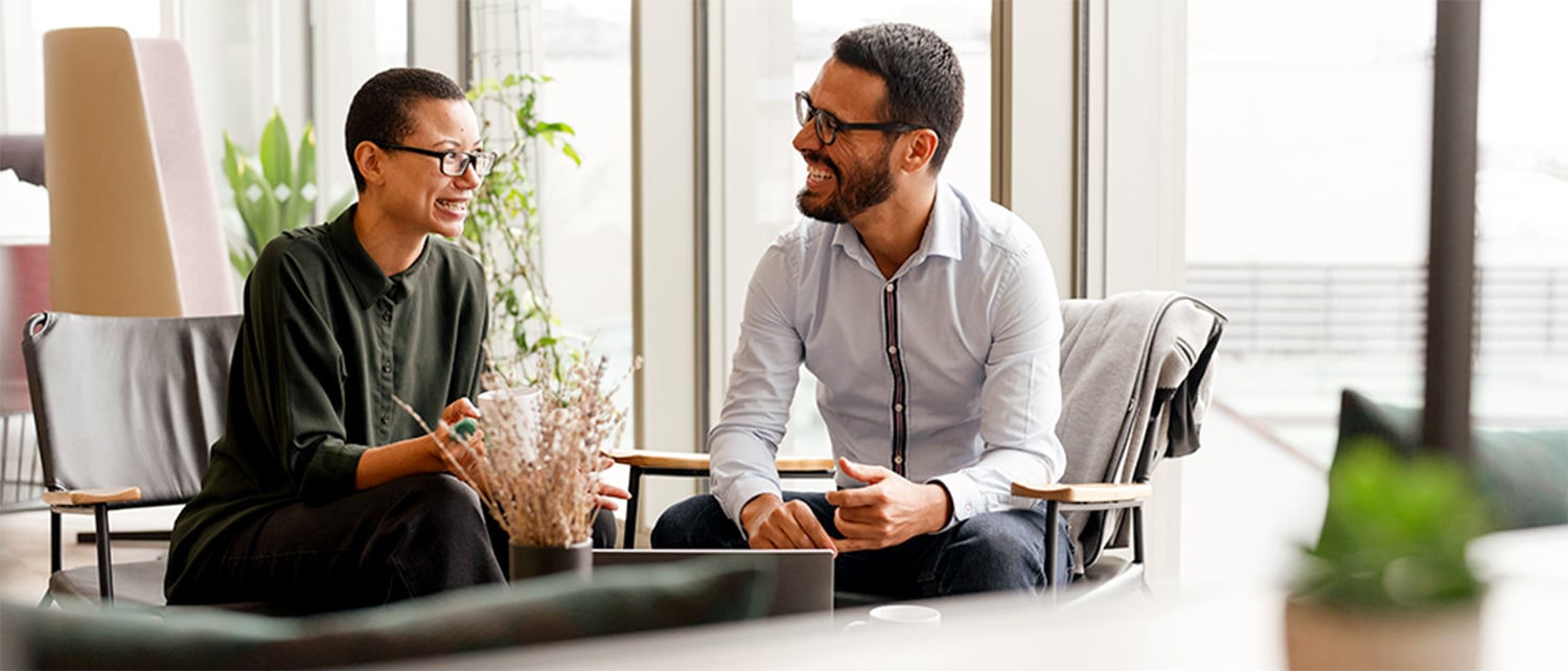 Two professionals sit in a bright modern office, smiling and engaged in conversation. One person gestures while speaking as the other listens attentively. Large windows, plants, and comfortable seating create a relaxed and collaborative workplace environment focused on teamwork and connection.