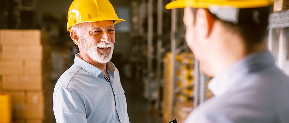 An older warehouse worker wearing a yellow hard hat and light blue shirt smiles while talking to a colleague in a storage aisle. The second worker is slightly out of focus in the foreground, with shelves and stacked boxes in the background, suggesting a friendly, collaborative work environment.