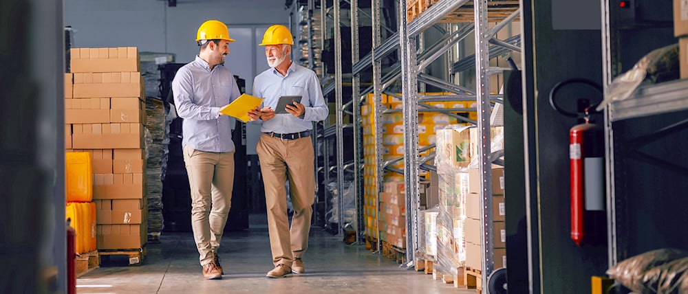 Two warehouse workers wearing yellow hard hats walk side by side through an aisle lined with shelves and stacked boxes. One holds a clipboard while the other uses a tablet, as they talk and review inventory in a well-organized industrial storage environment.