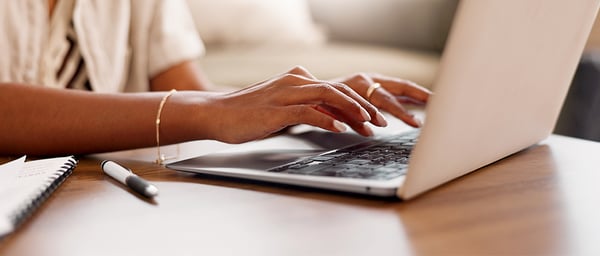 Close-up of hands typing on a laptop keyboard at a wooden desk in a warm, softly lit setting. A notebook and pen rest nearby, suggesting focused remote work, studying, or digital collaboration in a comfortable and organized workspace.