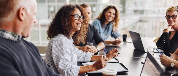 A diverse group of professionals sits around a conference table in a bright modern office, smiling and engaged in discussion. Laptops, notebooks, and coffee cups are visible, creating a collaborative atmosphere that suggests teamwork, brainstorming, or a productive business meeting.