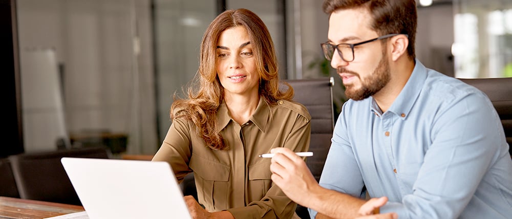 Two colleagues collaborating at a desk, reviewing content on a laptop while discussing ideas in an office setting.