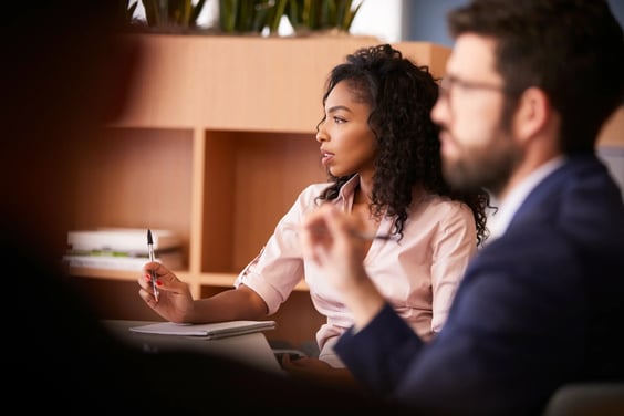 Woman taking notes with a pen while listening during a meeting, with a colleague blurred in the foreground.