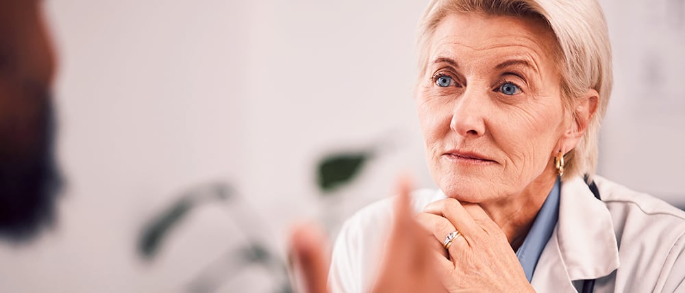 Medical professional in a white lab coat speaking during a healthcare consultation, with another person partially visible in the foreground.